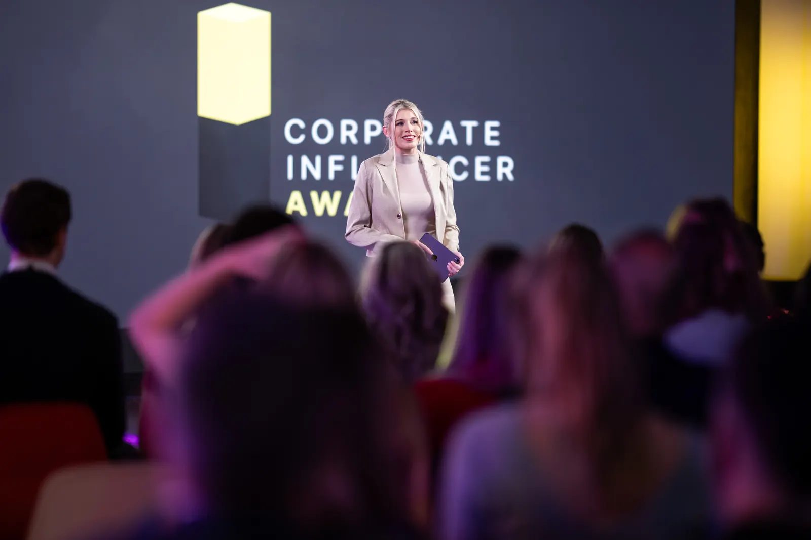 Corporate Influencer Award: Moderatorin auf der Bühne vor der Award-Wand, Publikum im Vordergrund
